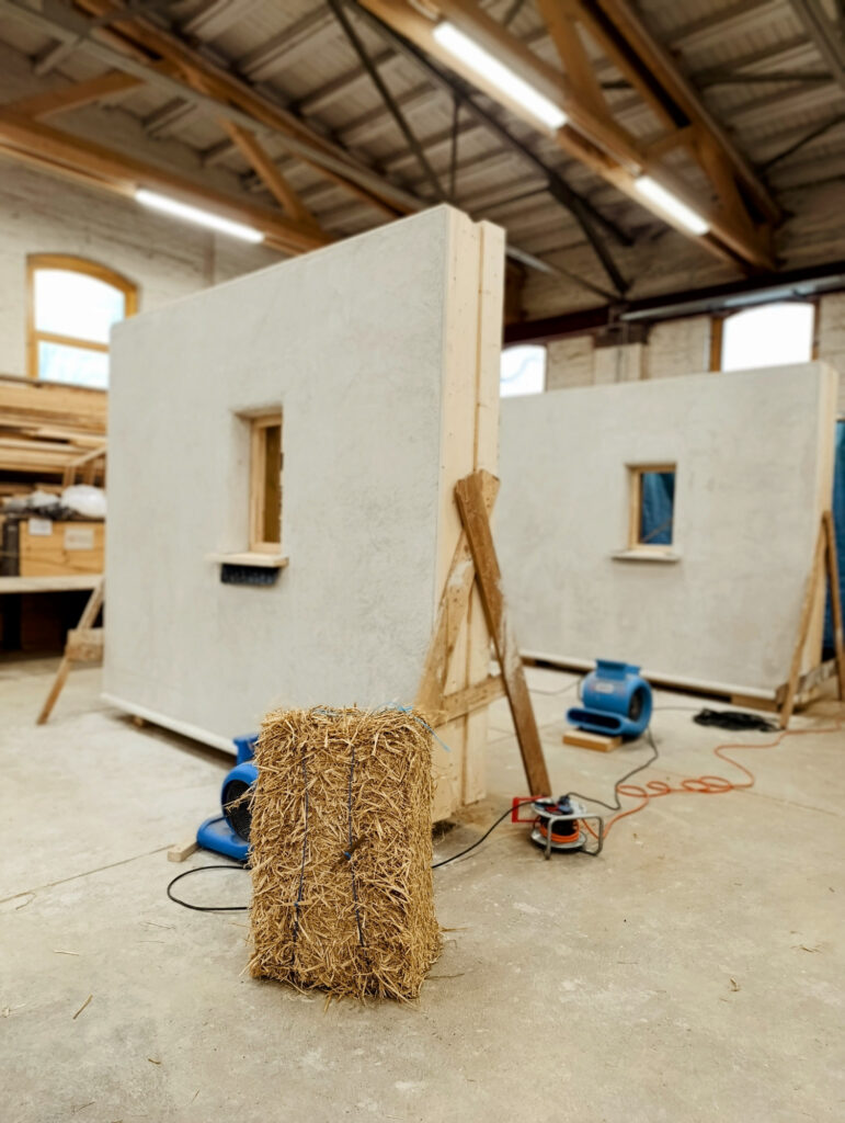 Baustroh-Ballen vor Strohelementen die mit Kalk verputzt sind.